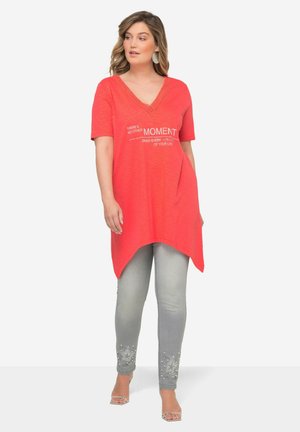 T-shirt tunica corallo con scollatura a V e maniche corte, caratterizzata da testo stampato bianco. Indossata con jeans grigio chiaro, impreziositi da stelle all'orlo.