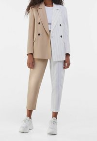 Béžovo-bílý splývavý blazer s černými knoflíky, kombinovaný s upnutými kalhotami; bílé kalhoty mají jemný proužek. Bílé tenisky doplňují celý vzhled.