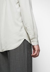 Camicia a maniche lunghe color beige chiaro con orlo curvo, polsini abbottonati e texture morbida, abbinata a pantaloni grigi con motivo.