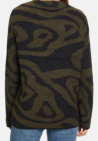 Pull en maille à motif abstrait vert foncé et noir porté par une personne vue de dos, au-dessus d'un jean bleu.