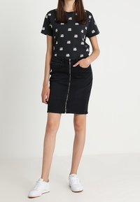 Jupe en denim noir avec une fermeture éclair à l'avant, associée à un tee-shirt noir à manches courtes avec un motif graphique blanc. Des baskets blanches viennent parfaire la tenue.