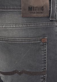 Jean en denim gris avec une étiquette en cuir portant l'inscription "MUSTANG True Denim". Présente des coutures décoratives et un petit élément en métal marqué.