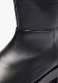 Bota de cuero negro hasta la rodilla con textura suave, punta redondeada y un sutil detalle de costura. Presenta un tacón grueso y un diseño minimalista.