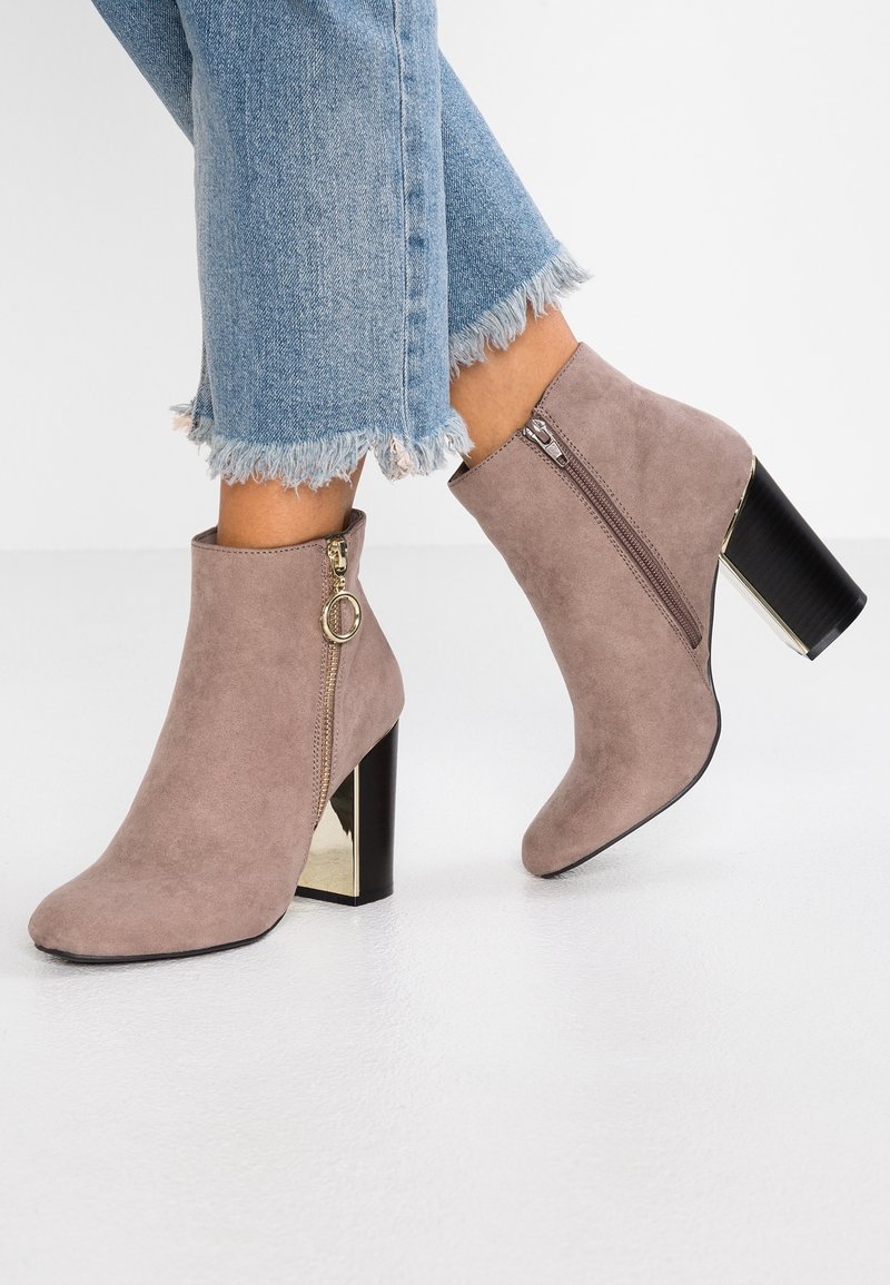 Bottines en daim taupe avec talons carrés noirs et fermetures éclair dorées, portées avec un jean bleu à ourlets effilochés sur un sol blanc.