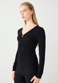 Schwarzes Langarmoberteil mit V-Ausschnitt, featuring frontale Raffung und einem goldenen Ringakzent in der Mitte. Hergestellt aus geripptem Material.