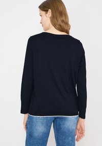 Femme aux cheveux châtain clair portant un haut noir uni à manches longues et un jean bleu, vue de dos sur fond blanc.