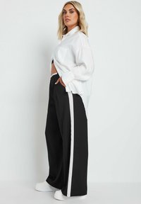 Biała, oversizowa koszula z kieszenią, w zestawieniu z czarnymi spodniami o szerokich nogawkach z białymi paskami po bokach. Modelka ma na sobie białe sneakersy.