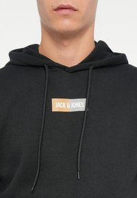 Felpa nera con cappuccio e coulisse regolabili, caratterizzata da un logo rettangolare arancione e grigio con la scritta "JACK & JONES." Tessuto morbido e texture.