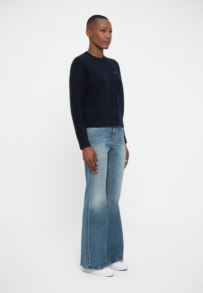 Navy cable-knit sweater with ribbed hem, paired with light blue flared jeans. White sneakers complete the outfit.