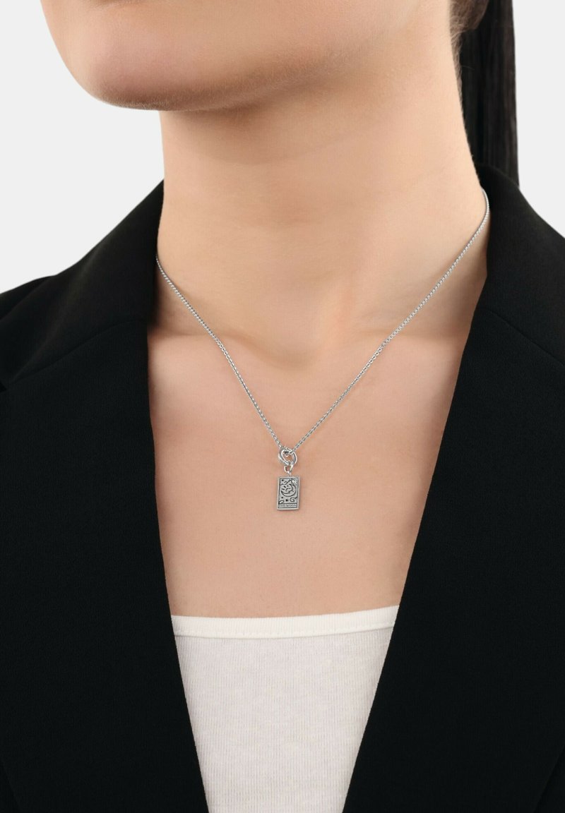 Collana in argento con un piccolo ciondolo rettangolare caratterizzato da un design astratto, indossata con un blazer nero e una blusa bianca.