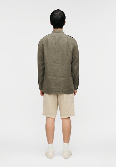 Πουκάμισο από λινο ύφασμα χρώματος ελιάς με μανσέτες με κουμπιά, φορεμένο πάνω από μπεζ σορτς. Το πουκάμισο έχει χαλαρή σχεδίαση με ομαλή υφή. Λευκά αθλητικά παπούτσια ολοκληρώνουν την εμφάνιση.
