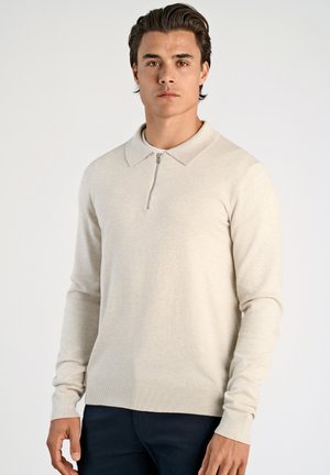 Suéter de pullover beige con cuello con cremallera, hecho de una tela suave. Presenta mangas largas y puños acanalados, usado sobre una camisa blanca.