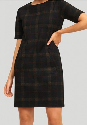 Donna che indossa un vestito a quadri nero e marrone, lungo al ginocchio con maniche corte, una mano sui fianchi, in piedi davanti a uno sfondo semplice.