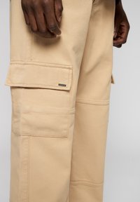 Beige cargobyxor i ett mjukt, strukturerat tyg. Funktioner inkluderar stora sidofickor, förstärkt sömnad och en liten metallisk logotypdetalj.