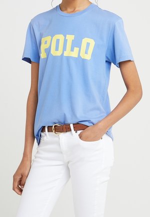 Persoon draagt een lichtblauw T-shirt met gele "POLO" tekst, ingestopt in een witte broek met een bruine riem, hand in de zak.