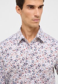 Uomo che indossa una camicia bianca con bottoni e un motivo floreale multicolore, colletto aperto, che guarda leggermente verso destra su uno sfondo semplice.