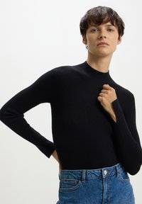 Maglia nera a maniche lunghe con collo alto. Il tessuto ha un aspetto testurizzato. Abbinata a jeans di denim blu con tasche classiche.