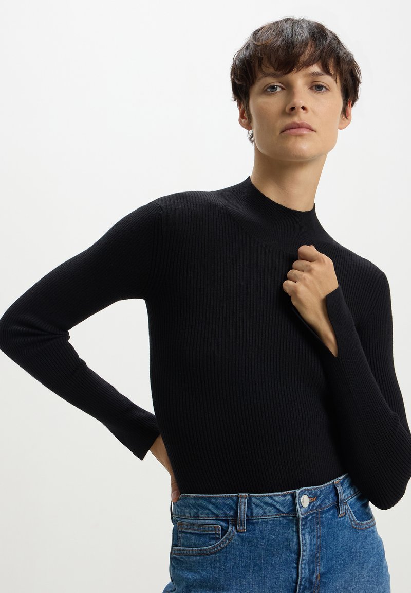 Maglia nera a maniche lunghe con collo alto. Il tessuto ha un aspetto testurizzato. Abbinata a jeans di denim blu con tasche classiche.