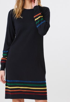 Nainen, joka pukeutunut mustaan pitkähihaiseen mekkoon, jossa on vaakasuoria, sinisiä, vihreitä, keltaisia, oransseja ja punaisia kimaltavia raitoja hihoissa ja helmassa.