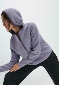 Lila Fleece-Hoodie mit einem halben Reißverschluss vorne, elastischen Bündchen und einer gefütterten Kapuze. Getragen mit schwarzen Hosen, die eine weiche, strukturierte Oberfläche aufweisen.