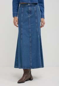 Jupe longue en denim bleu avec des coutures verticales, portée avec des bottes de cowboy en cuir marron et un blouson en denim assorti.