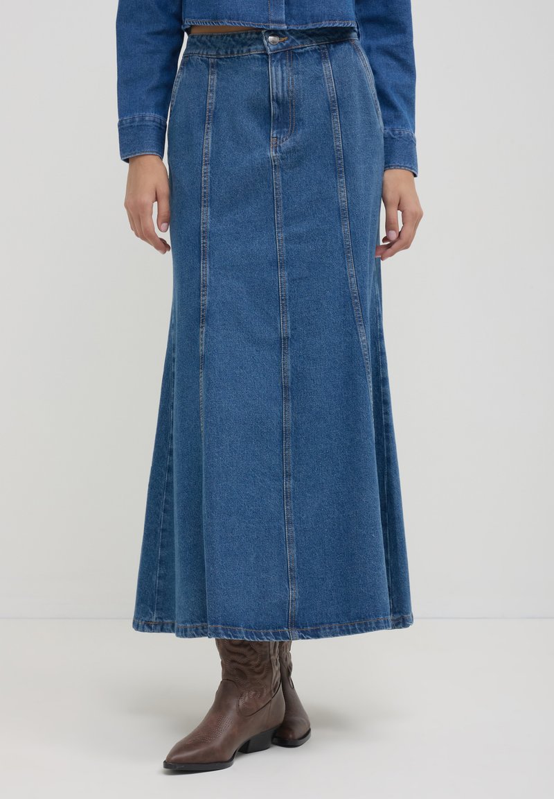 Jupe longue en denim bleu avec des coutures verticales, portée avec des bottes de cowboy en cuir marron et un blouson en denim assorti.