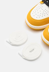 Sárga sportcipők fehér perforált orral, fehér fűzőkkel és texturált gumitalppal, két feltekert fehér fűzővel egy fehér háttéren.