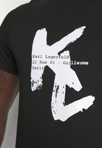 Black t-shirt featuring a large white brushstroke letter 'K' and text reading "Karl Lagerfeld 21 Rue St - Guillaume Paris." Smooth fabric texture.