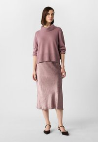 Lila Strickpullover mit Schalkragen und oversize Passform, kombiniert mit einem metallic rosa Midirock mit strukturiertem Finish. Braune Absatzschuhe.