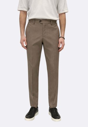 Uomo che indossa pantaloni sartoriali color marrone chiaro, una camicia bianca a maniche corte, scarpe da ginnastica nere con suole bianche e un braccialetto nero al polso destro.