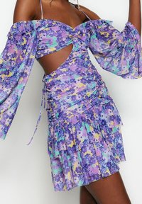 Lilla blomsterkjole med off-shoulder ærmer, udskæring, rynket tekstur og lagdelt nederdel med gule og grønne detaljer.