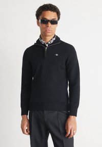 Hombre con suéter negro con cremallera, bufanda negra y blanca con estampado, pantalones negros y gafas de sol oscuras sobre un fondo blanco liso.