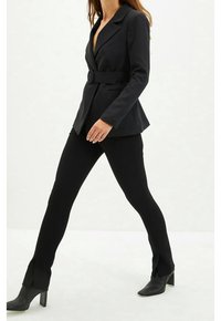 Musta räätälöity bleiseri kolmiomallisella kauluksella, vyötäröllä soljella ja kahdella etutaskulla; yhdistetty vartalonmyötäisiin mustiin housuihin ja korollisiin nilkkureihin.