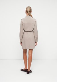 Beige cotton dress with long sleeves, belted waist, and buttoned cuffs, featuring a straight cut that falls above the knees. Worn with dark flats.