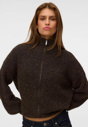 Jeune femme portant un pull zippé duveteux marron foncé, mains sur les hanches, sur un fond clair uni.