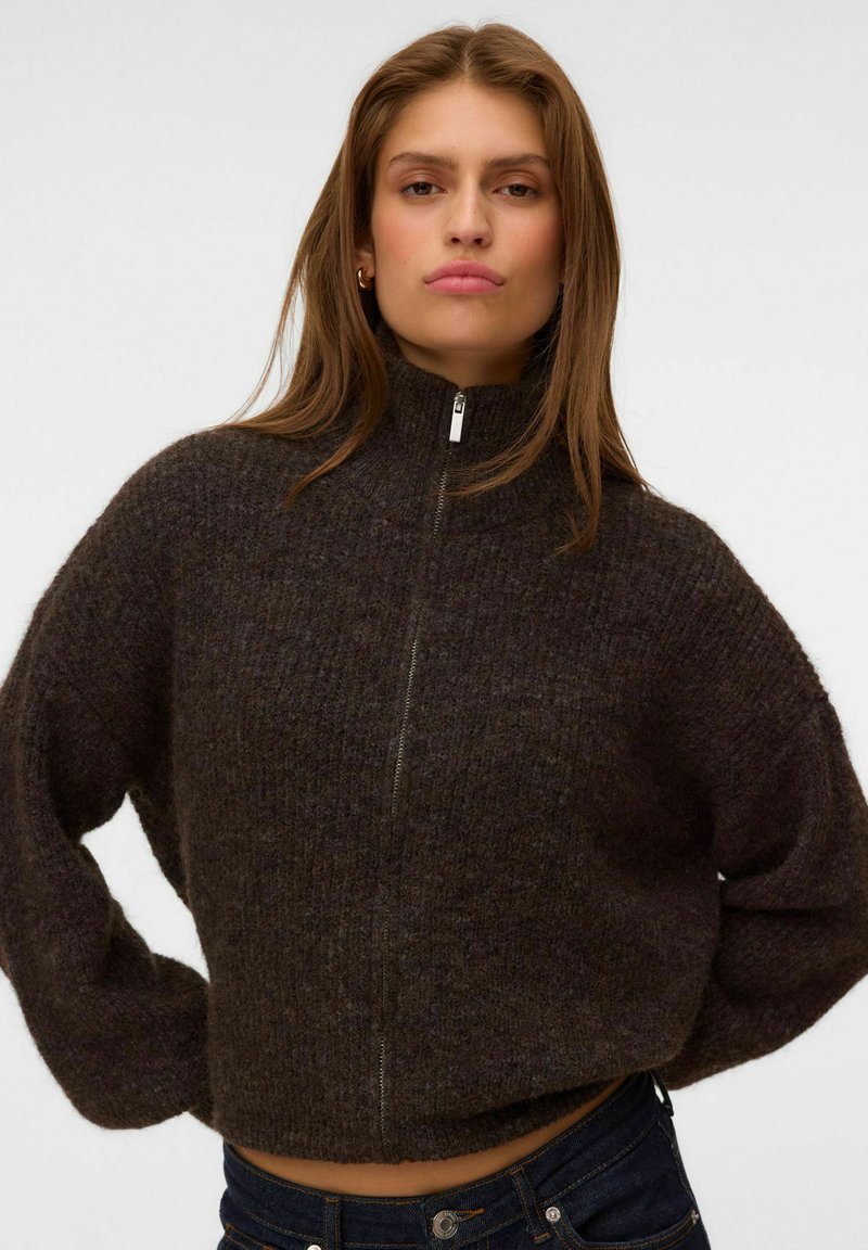 Jeune femme portant un pull zippé duveteux marron foncé, mains sur les hanches, sur un fond clair uni.