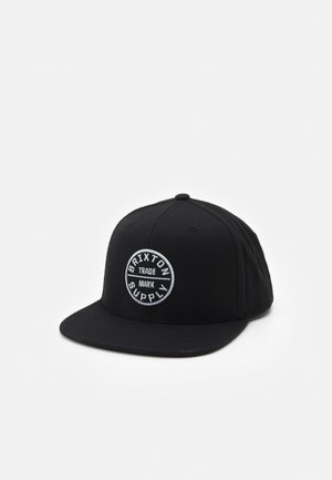 Schwarze flache Kappe mit weißem rundem Logoaufnäher mit der Aufschrift "Brixton Supply Trade Mark" vorne, dargestellt auf weißem Hintergrund.