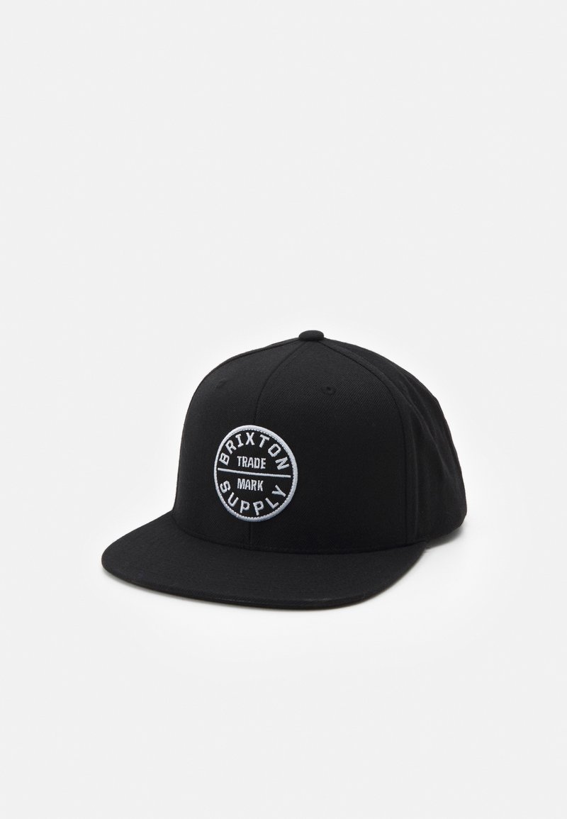 Casquette noire à visière plate avec un écusson logo circulaire blanc portant l'inscription "Brixton Supply Trade Mark" sur le devant, affichée sur un fond blanc.