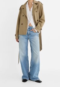 Khaki trench coat with large buttons over a white shirt, paired with light-wash wide-leg jeans featuring distressed details and black shoes.