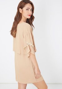 Joven mujer con cabello largo castaño, vistiendo un vestido beige suelto con un detalle de lazo floral en la manga, posando contra un fondo blanco.