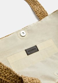Geflochtener Strohtäschchen mit einem beigen Innenfutter aus Stoff, ausgestattet mit einem Druckknopf, einem Lederetikett und einer Tasche im Lining.