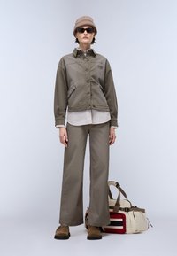 Ελαιοπράσινο κοντό πανωφόρι με κουμπιά και τσέπες, συνδυασμένο με ριγέ ανοιχτό πουκάμισο και φαρδιά ελαιοπράσινα παντελόνια. Καφέ παπούτσια και πολύχρωμη τσάντα.