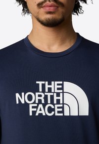 Dunkelblaues T-Shirt mit Rundhalsausschnitt. Mit großem weißen Logotext "THE NORTH FACE" und einem geschwungenen grafischen Design.