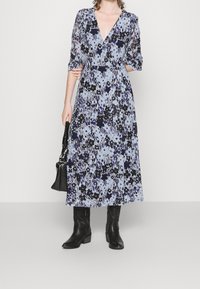 Długa sukienka w kwiatowy nadruk w jasnoniebieskim kolorze z czarnymi i fioletowymi akcentami. Dekolt w serek, bufiaste rękawy i swobodny krój z bocznymi rozcięciami.