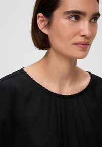 Zwarte blouse met een gegolfde halslijn, met verticale plooien en een gladde stofstructuur. Close-up van de halslijn en schoudergebied.