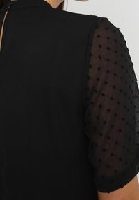 Top negro con un cuerpo de tela suave y mangas abullonadas transparentes que presentan pequeños lunares en relieve. Cierre de cremallera en la parte posterior visible.