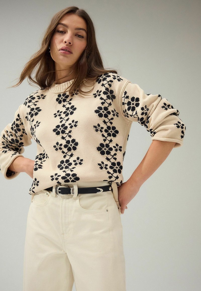 Pull en maille beige avec un motif floral noir, col rond côtelé et manches retroussées, associé à un pantalon crème et une ceinture noire.