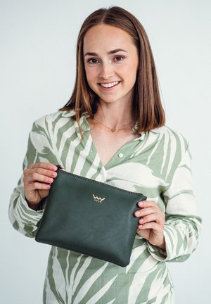Green leather clutch bag with gold hardware, zip closure, and logo. Model holds the bag in front of a light background, wearing a leaf-patterned shirt.
