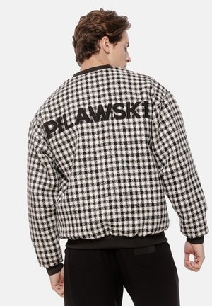 Mężczyzna ubrany w czarno-białą kurtkę w kratę z napisem "PILAWSKI" na plecach, w połączeniu z czarnymi spodniami, stojący tyłem.
