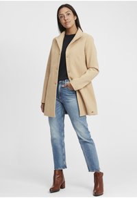 Helle beige Wolljacke mit einem Reverskragen, Knopfverschluss und taillierter Passform. Getragen über einem schwarzen Oberteil, kombiniert mit blauen Jeans und braunen Stiefeln mit Absatz.